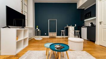 Un espacio habitable moderno con una pared azul oscuro de acento, un mueble blanco para entretenimiento, un área de cocina con microondas y una zona de estar con una mesa redonda y taburetes.