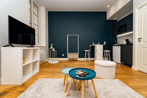 Un espace de vie moderne avec un mur d’accent bleu foncé, un meuble de rangement blanc pour l’équipement audiovisuel, un coin cuisine doté d’un micro-ondes et un espace détente avec une table ronde et des tabourets.