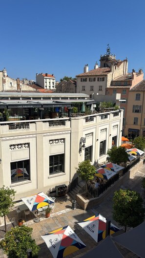 Un immeuble dont le rez-de-chaussée abrite un café et un restaurant, avec des tables à l’extérieur protégées par des parasols, et un ciel bleu dégagé.
