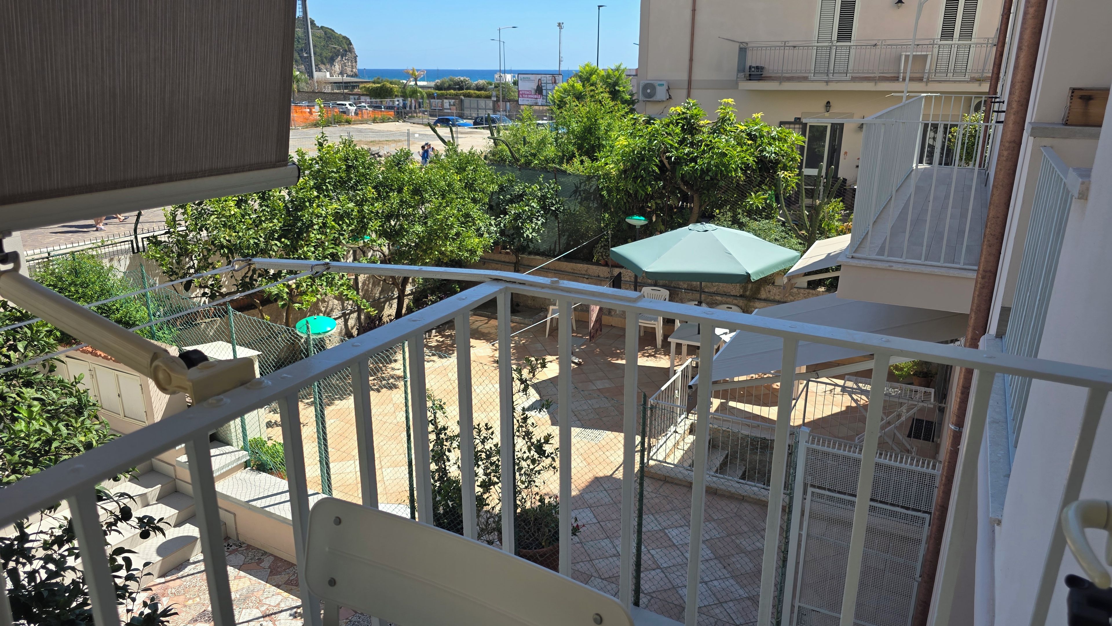 A balcony with a view of a courtyard, outdoor seating, and a distant view of the sea.