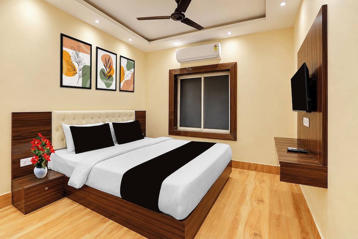 A hotel room with a bed, a nightstand, a vase of flowers, a ceiling fan, an air conditioning unit, a window, and a television.