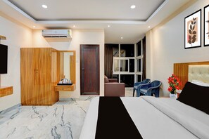 A modern hotel room with a large bed, a wooden wardrobe, a desk with a chair, a window with curtains, and a TV on the wall.