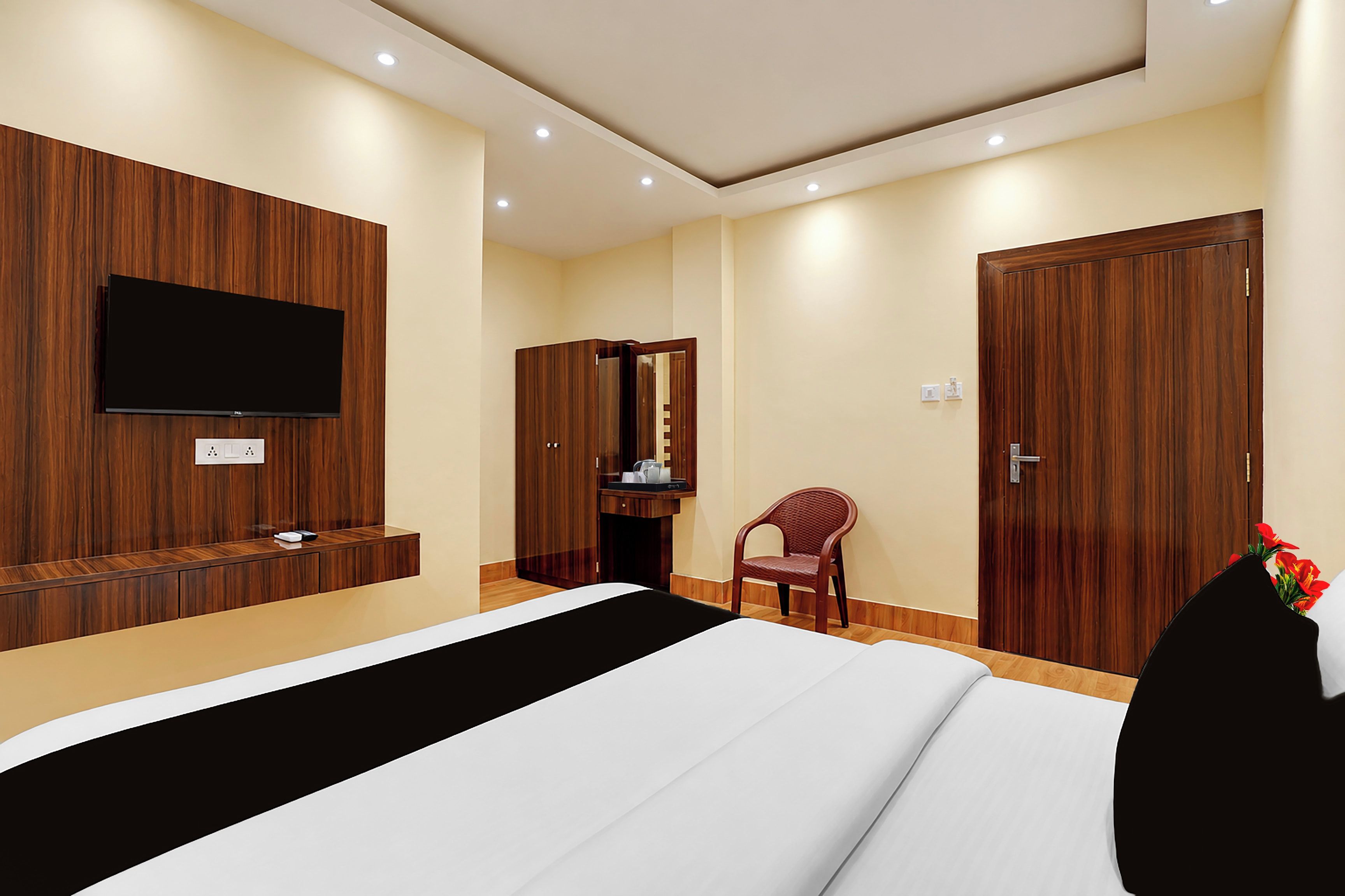 A hotel room with a bed, a TV mounted on the wall, a wooden wardrobe, a desk with a chair, and a door.