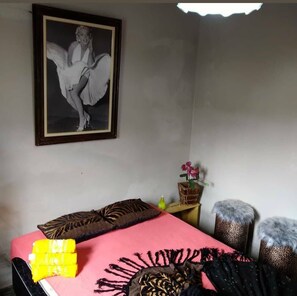 A bedroom with a bed, pillows, a framed picture on the wall, a small table with a vase, and two ottomans.