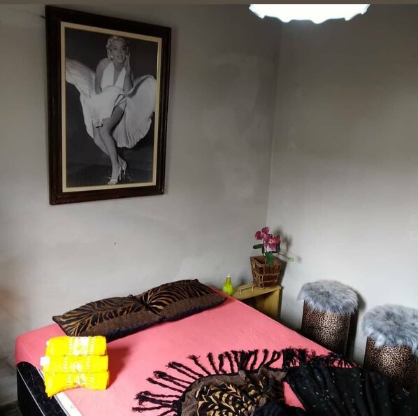 A bedroom with a bed, pillows, a framed picture on the wall, a small table with a vase, and two ottomans.