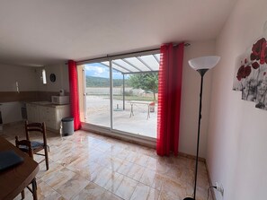 Une pièce comprenant une table à manger, des chaises, un coin cuisine, une grande porte-fenêtre coulissante donnant sur une terrasse extérieure et un lampadaire.