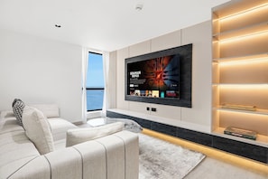 A modern living room with a large flat-screen TV mounted on the wall, a sofa, and a bookshelf with illuminated shelves. - FIRST CLASS | 3BR | Near JBR Beach | Ain Views (JBR)