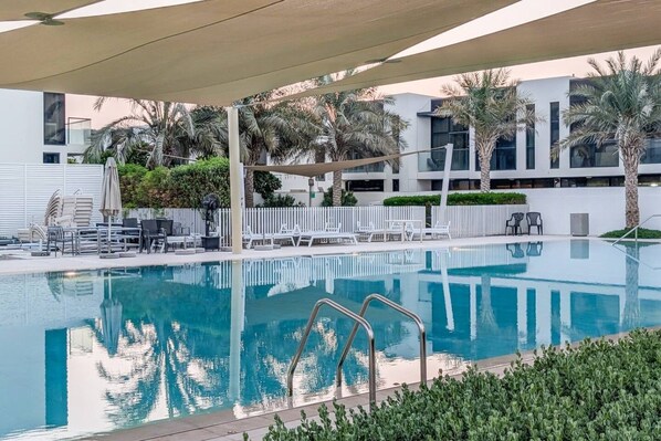 A swimming pool with lounge chairs and umbrellas, surrounded by palm trees and modern buildings.