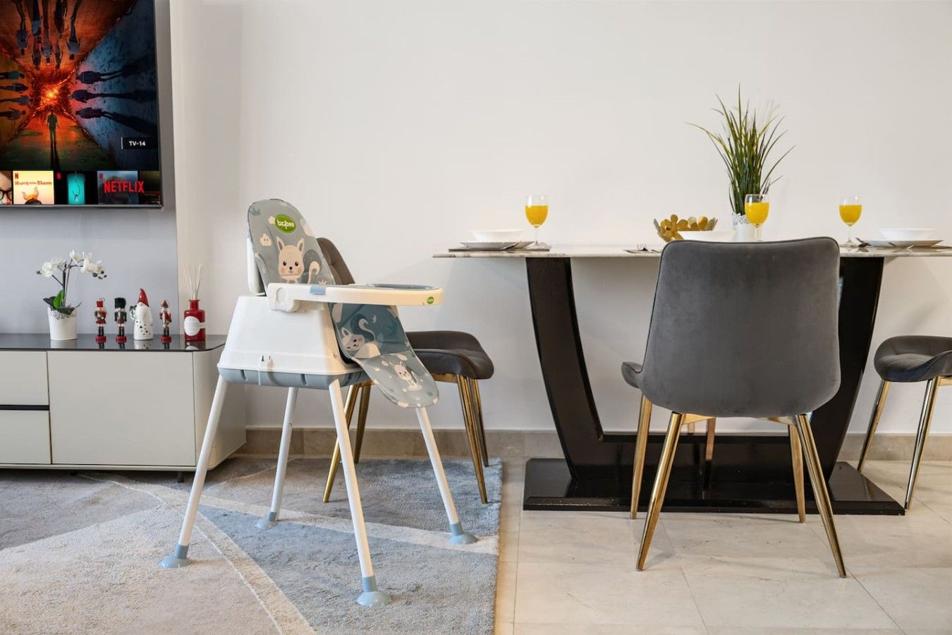A modern dining area with a high chair, a dining table with chairs, and a TV mounted on the wall displaying a movie.