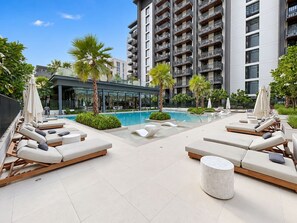 A rooftop pool area with sun loungers, palm trees, and a modern building in the background. - FIRST CLASS | 1BR | Lavish Living | Near Downtown (Sobha Hartland)
