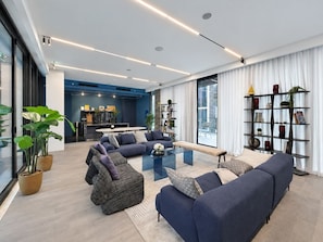 A modern living room with a large sectional sofa, a glass coffee table, and a dining area in the background. - FIRST CLASS | 1BR | Lavish Living | Near Downtown (Sobha Hartland)