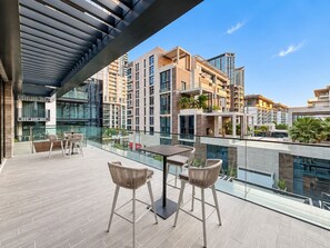 Una terraza moderna al aire libre con barandilla de vidrio, una mesa con sillas y vistas a edificios altos circundantes.