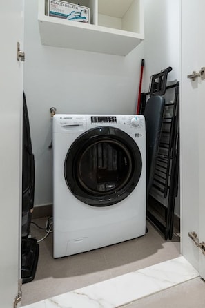 Eine kompakte, weiße Waschmaschine mit Frontlader in einem kleinen Waschraum mit einem Regal darüber und einem Faltstuhl zur Rechten.