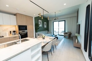 A modern kitchen with a dining area, a living room with a sofa and a coffee table, and a large window providing natural light. - FIRST CLASS | 1BR| Luxe Urban Living (Sobha Hartland)
