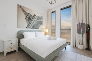 A hotel room with a large bed, a nightstand, a lamp, a painting of the Eiffel Tower, and a view of the landscape through the window. - FIRST CLASS | 1BR | Warm & Stylish Retreat (Dubai Production City)