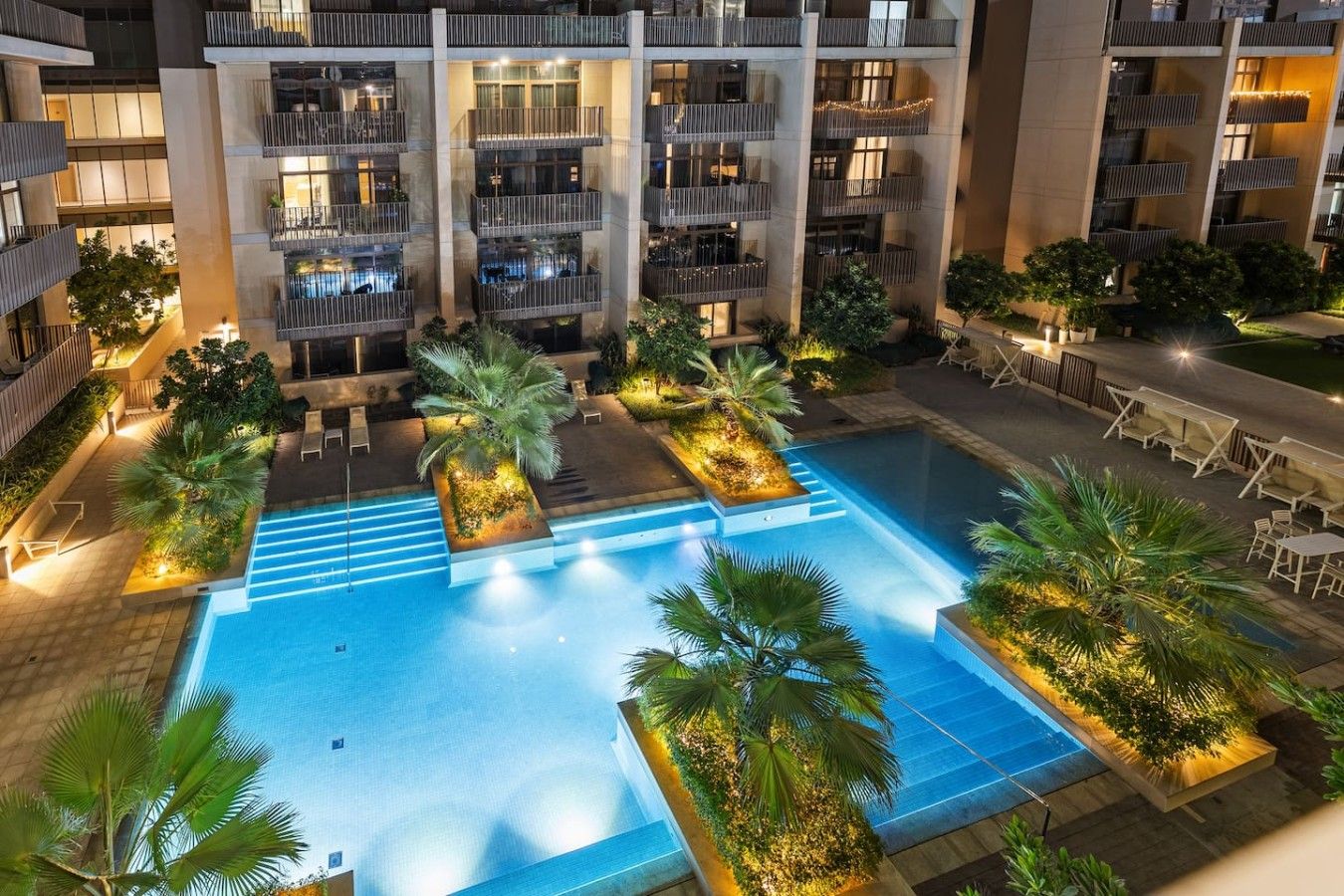 Un hotel de varios pisos con piscina central rodeada de mobiliario exterior y vegetación.