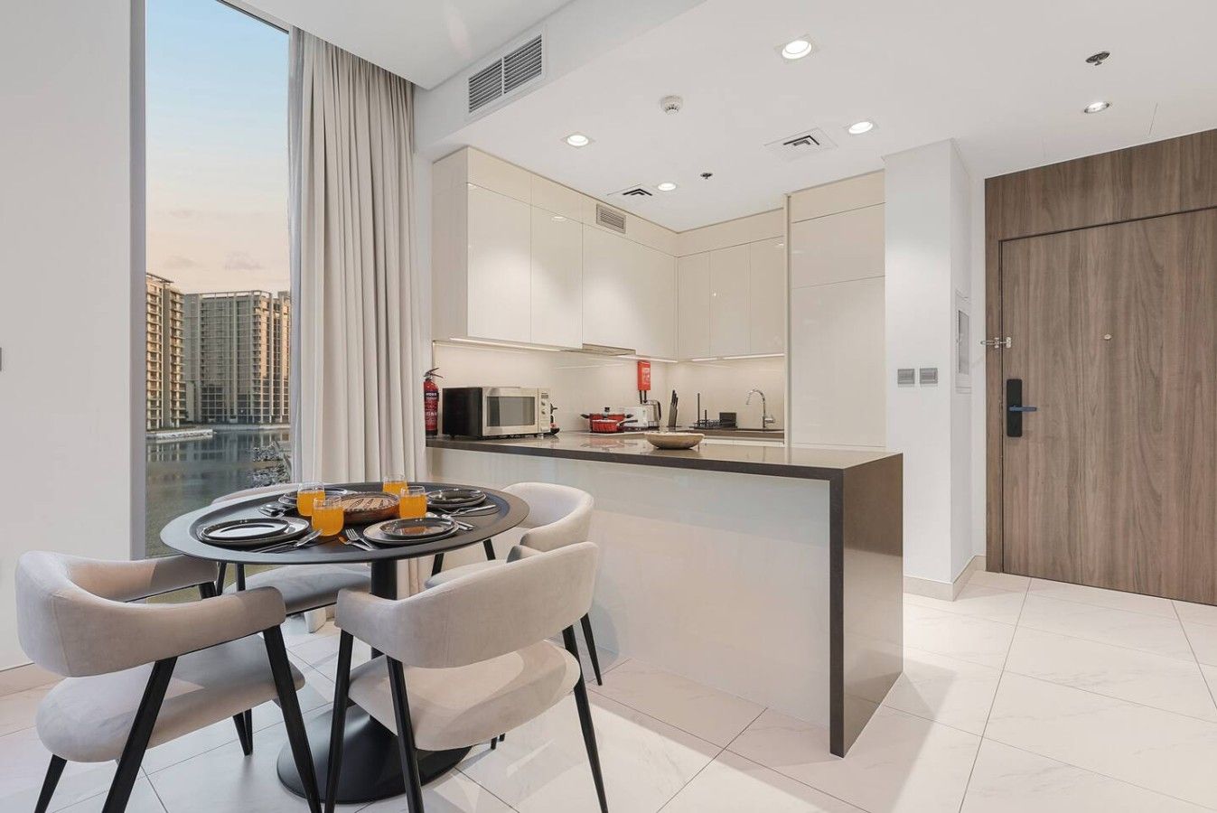 A modern kitchen with a dining area, large windows, and a view of high-rise buildings.