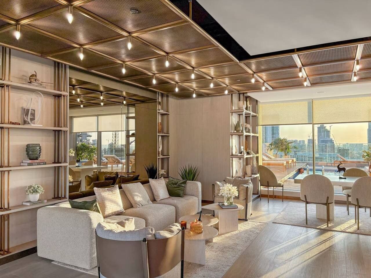 A modern living room with a sectional sofa, coffee table, and chairs. There are bookshelves and a large window with a city view.