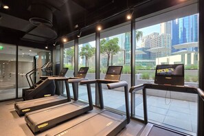 A fitness area with treadmills and a laptop on a table, large windows with a city view, and a staircase leading to an upper level. - FIRST CLASS | 2BR | Tranquil Canal View Haven (Business Bay)