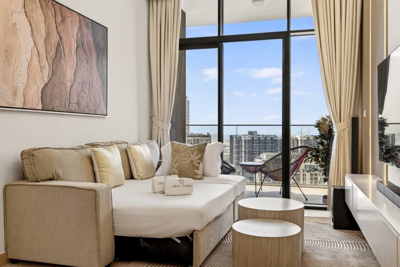 A modern hotel room with a large bed, a sofa, two side tables, and a view of the city through large windows.