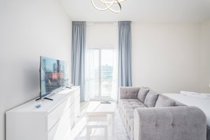 A modern living room with a flat-screen TV, a sofa, a white cabinet, and a view of the city through large windows. - FIRST CLASS | Studio | Lavish Cozy Haven (Dubai Sports City)