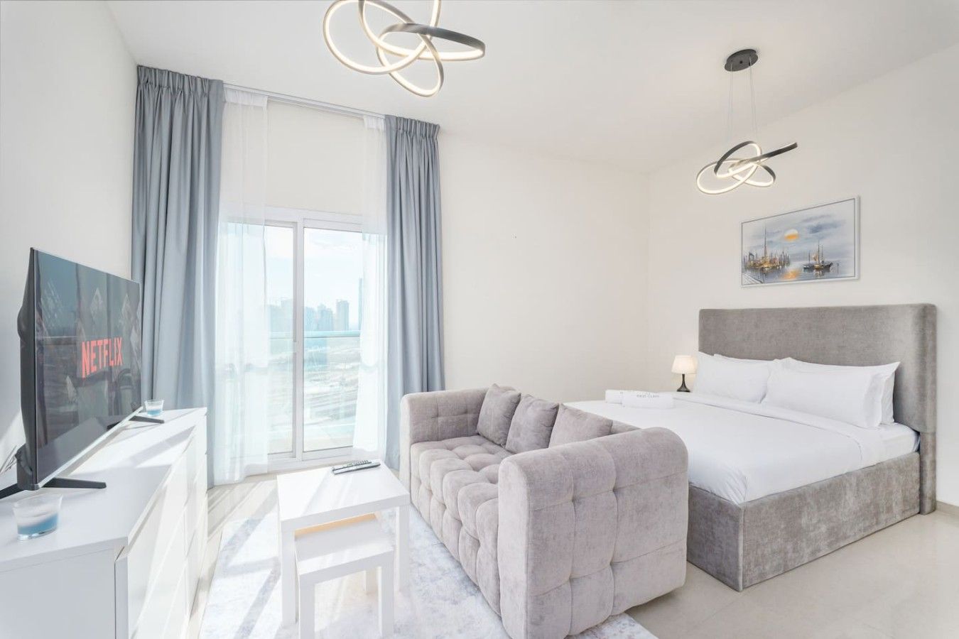 A modern hotel room with a bed, a sofa, a TV, and a large window with curtains.
