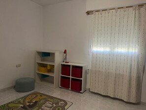 Ein Zimmer mit weißem Boden, einem bunten Teppich, einem weißen Schrank mit roten und gelben Boxen, einer roten Lampe und einem weißen Vorhang mit Muster.