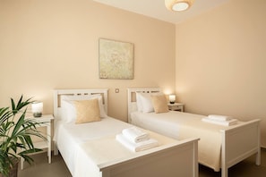 A hotel room with two beds, a bedside table with a lamp, a wall-mounted artwork, and a potted plant. - Elios Estate | 22-Guest Villa with Heated Pool (Marathi)