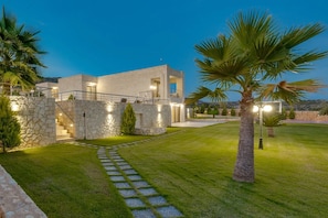 A modern house with a stone facade, a well-maintained lawn, and a palm tree. - Elios Estate | 22-Guest Villa with Heated Pool (Marathi)