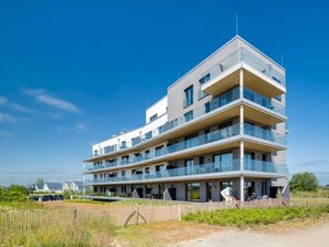 Ein mehrgeschossiges Wohngebäude mit Balkonen, einer flachen Dachterrasse und blauem Himmel.