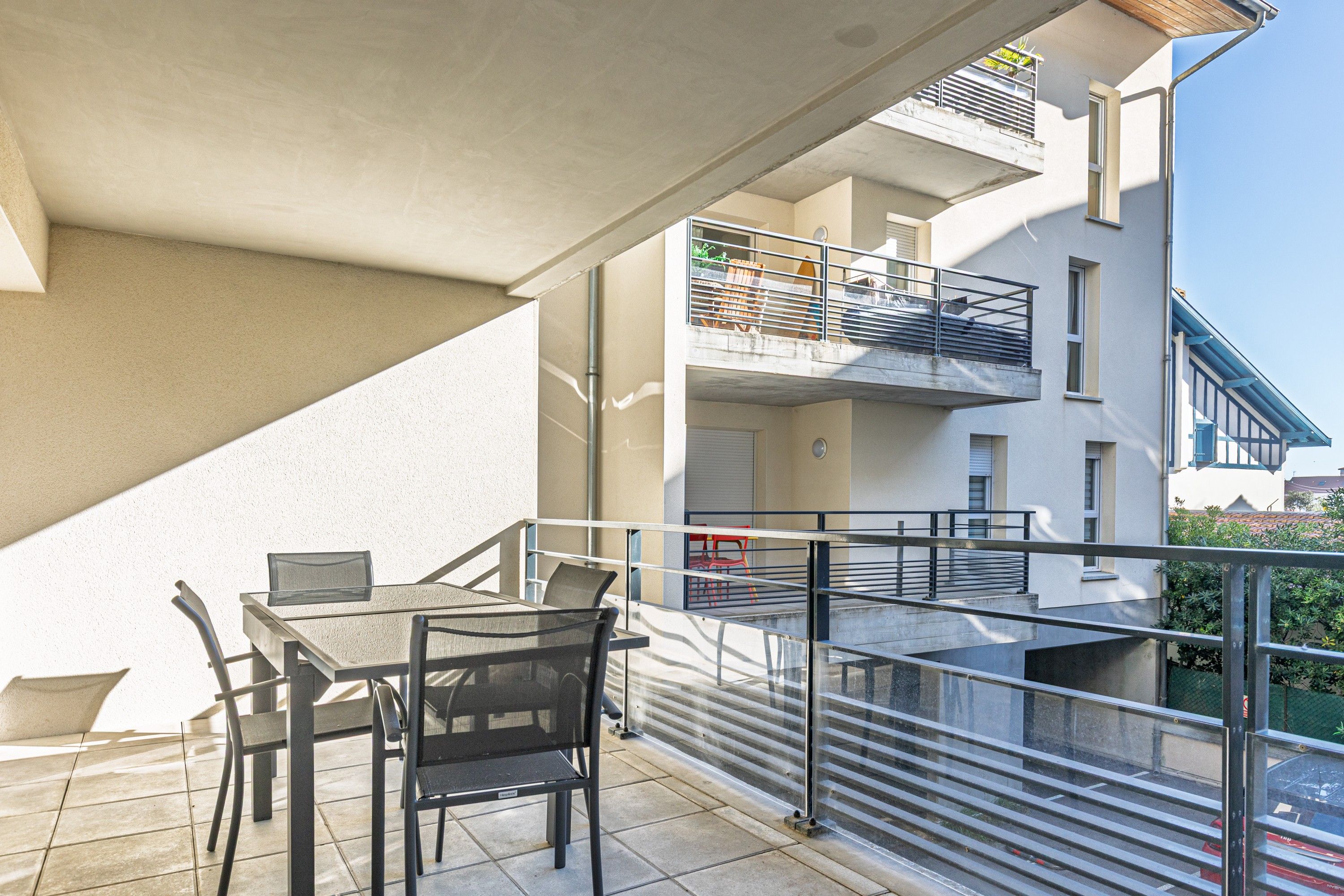 An outdoor balcony with a table and chairs, a metal railing, and a view of adjacent buildings.