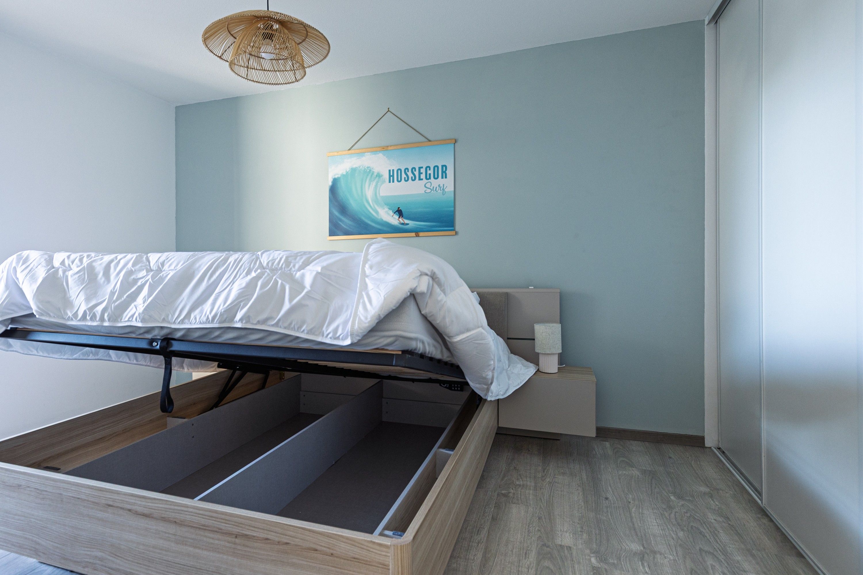 A bedroom with a bed, a nightstand, a wall-mounted lamp, and a poster of a surfer.