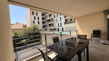 Un balcón con una mesa y sillas de vidrio, vistas a una piscina y un edificio con múltiples balcones al fondo.