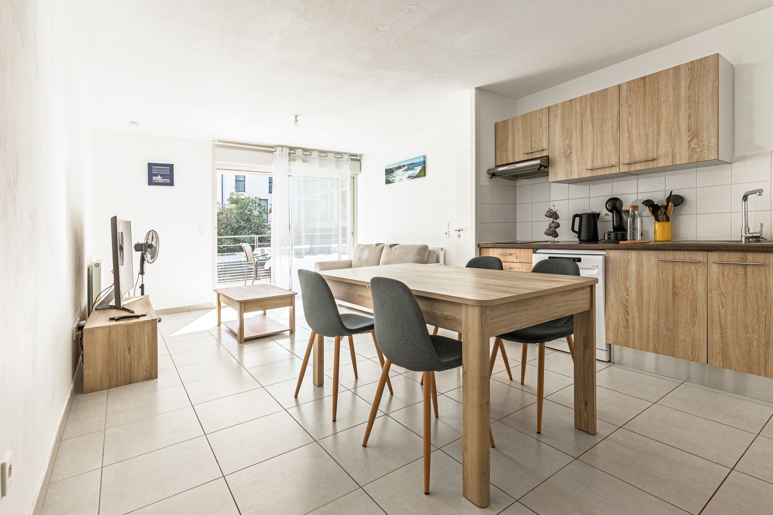 A modern kitchen with wooden cabinets, a dining table with chairs, and a view of the outdoors through a glass door.