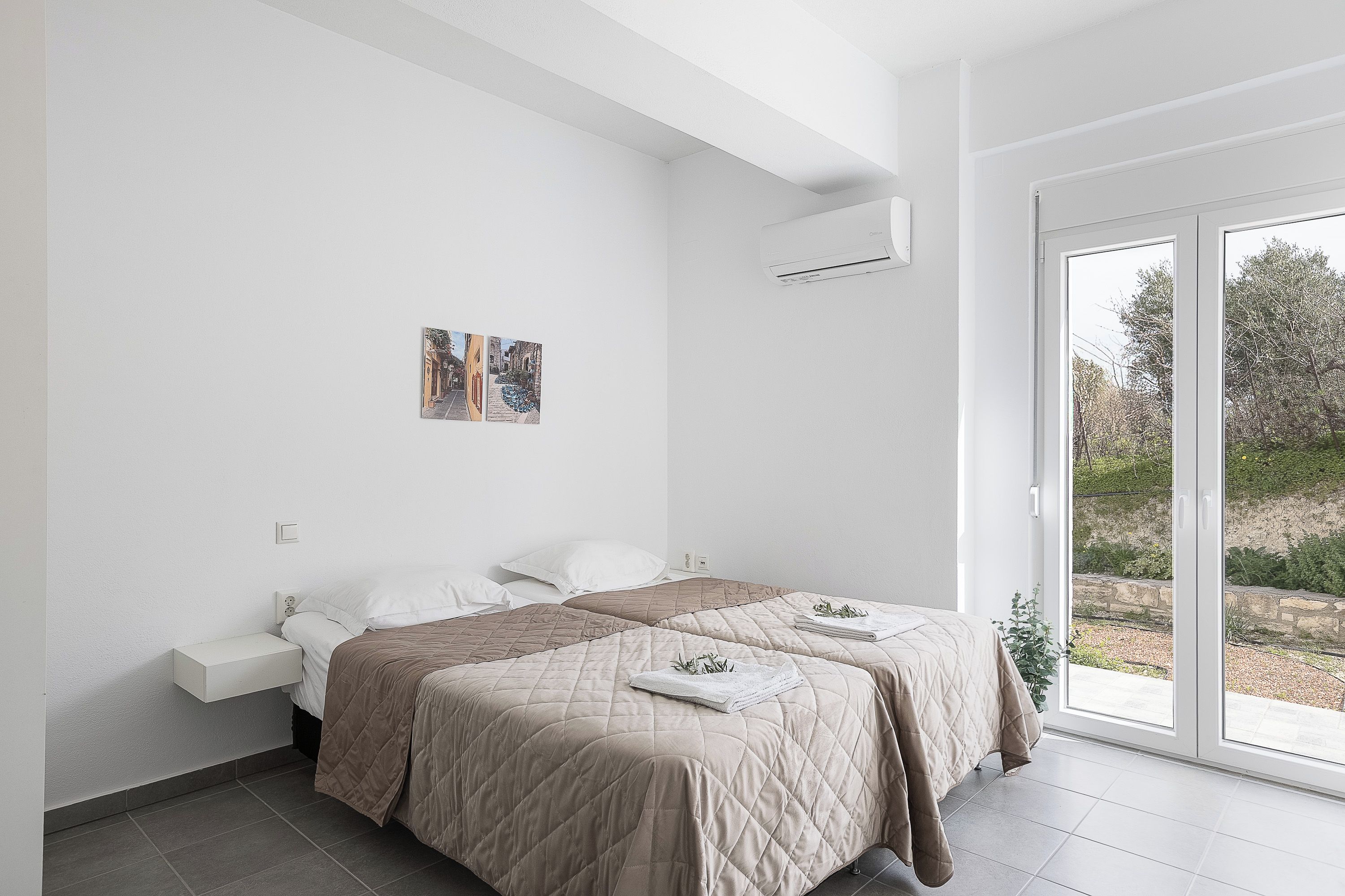 A modern bedroom with two beds, a white wall, a small artwork, and a large sliding glass door to the outdoors.