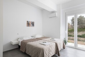 Une chambre moderne avec deux lits, un mur blanc, une petite œuvre d’art et une grande porte-fenêtre coulissante donnant sur l’extérieur.