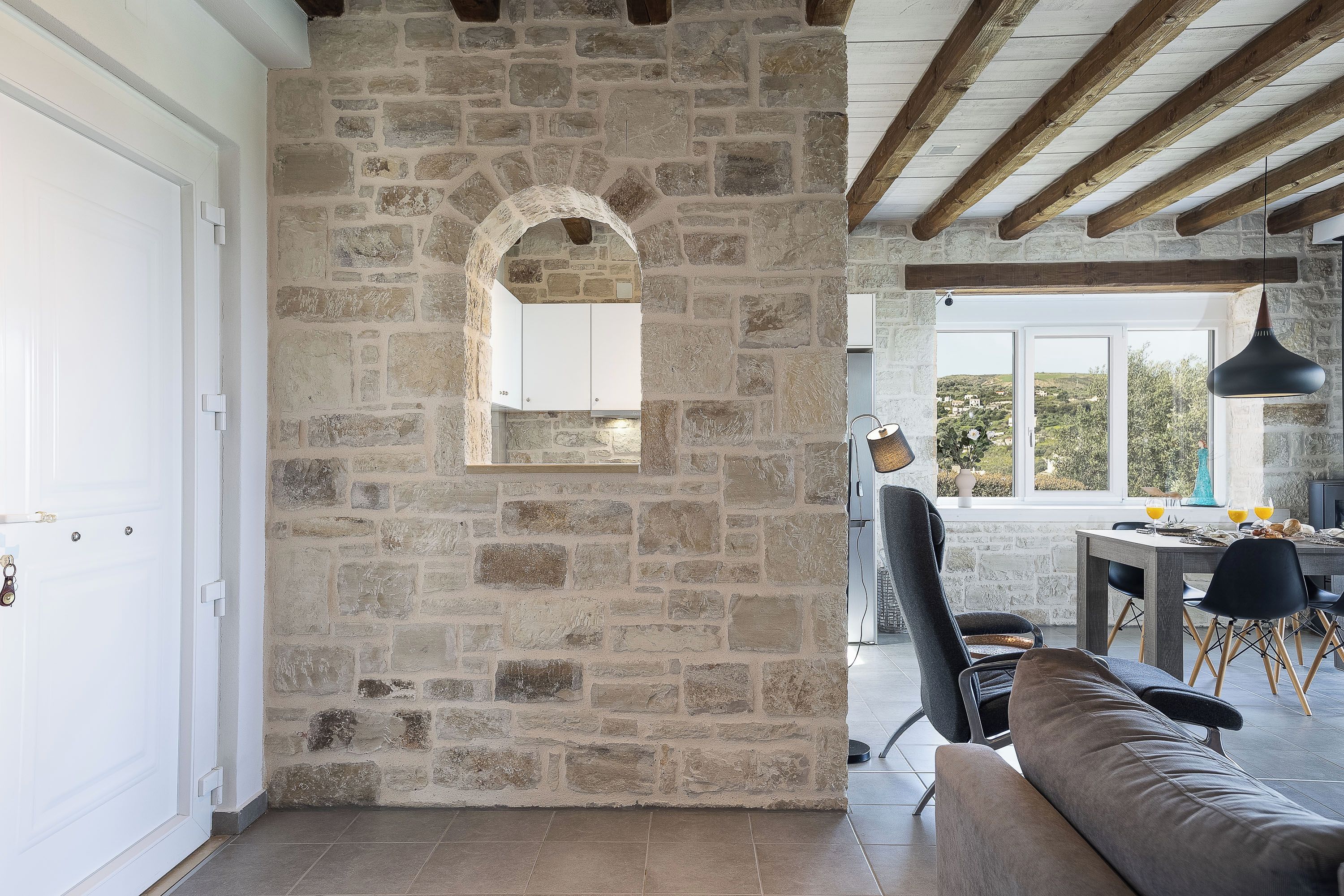 A stone-walled room with a wooden ceiling, a dining area with a table and chairs, and a view of the outdoors.