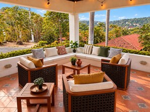 A covered outdoor seating area with a sofa, chairs, and tables, surrounded by lush greenery and a view of a residential area. - Villa 33 | Tropical Retreat at Villas Sol (Playa Hermosa)