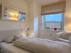 A bedroom with a bed, a chair, a radiator, a window with curtains, a lamp, and a mirror. - Holiday Apartment With 3 Bedrooms (Wyk auf Foehr)