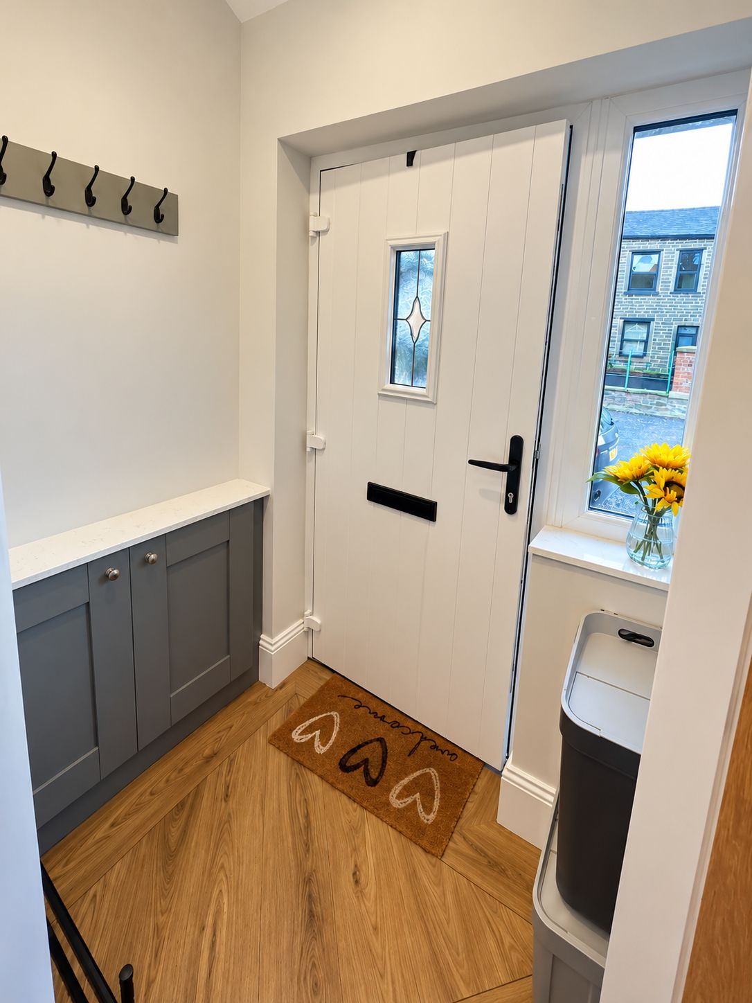 A hallway with a wooden floor, a white door with a glass window, a coat rack, and a trash bin.
