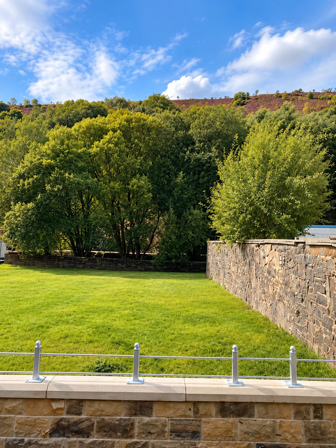 A grassy area with a stone wall and metal railings, likely part of a park or garden setting.