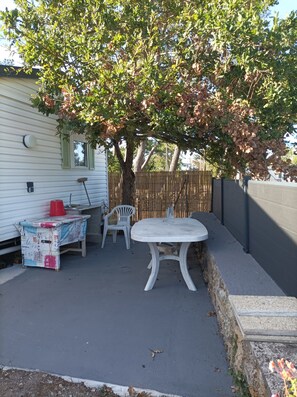 Een terras met een witte tafel en stoelen, een rode emmer, en een overdekte tafel met een tafelkleed met patroon.