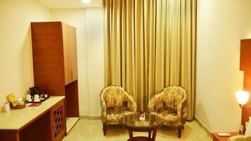 Habitación de hotel con un armario de madera, una pequeña mesa con superficie de vidrio, dos sillones estampados, una cortina y una lámpara de noche.