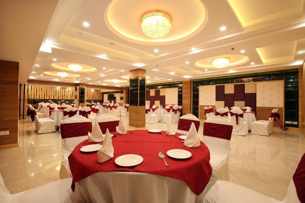 Un salón de banquetes con mesas redondas dispuestas para comer, sillas blancas con cintas rojas y una lámpara de araña moderna.