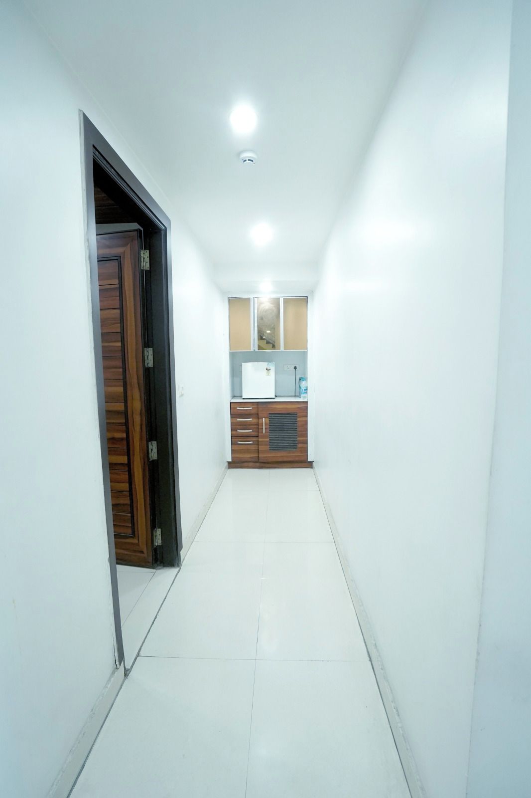 A narrow hallway with a wooden door on the left, a white tiled floor, and a kitchenette area with cabinets and a refrigerator in the distance.