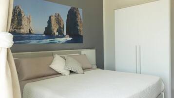 Un dormitorio con cama, almohadas, cabecero, armario y un cuadro de un paisaje costero.