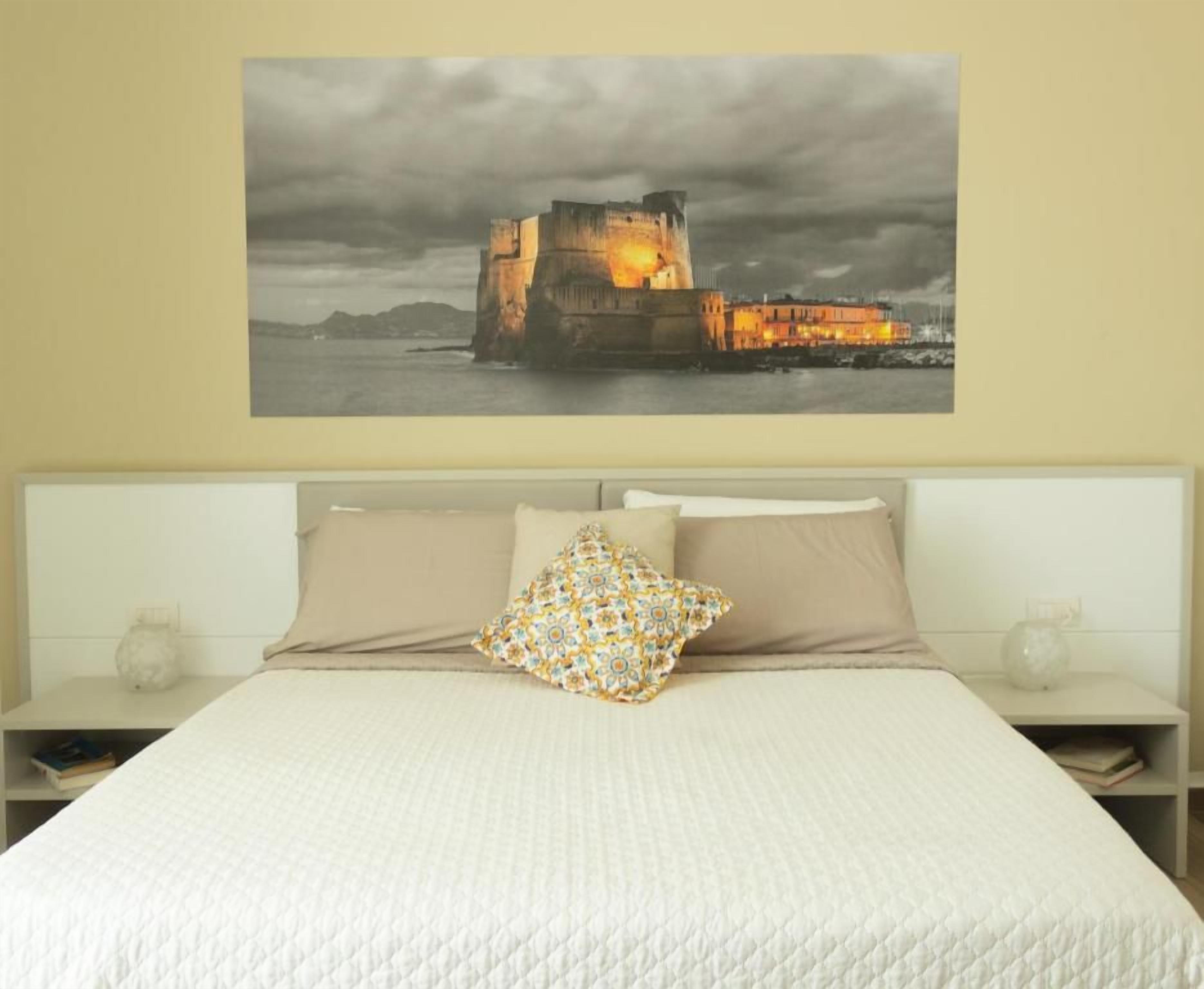 Un dormitorio con una cama grande, una mesita de noche, una lámpara de pared y un cuadro con un castillo.