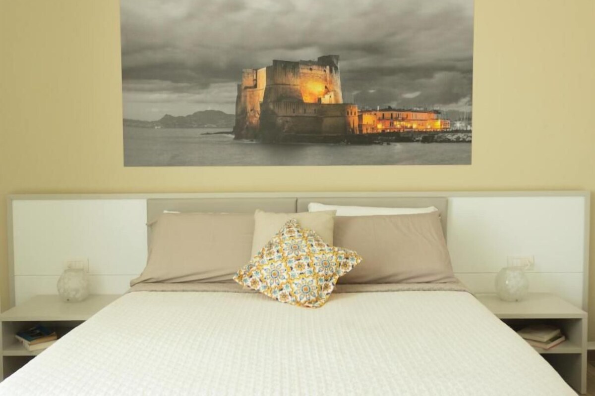 Una camera da letto con un letto grande, un comodino, una lampada a muro e un quadro di un castello.