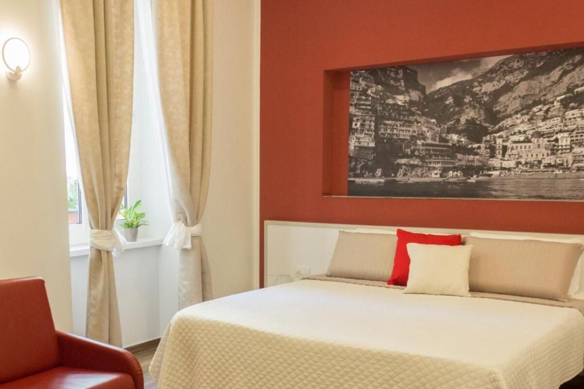 Camera d'albergo con un letto, una poltrona rossa, una finestra con tende e un quadro appeso al muro raffigurante una località costiera.
