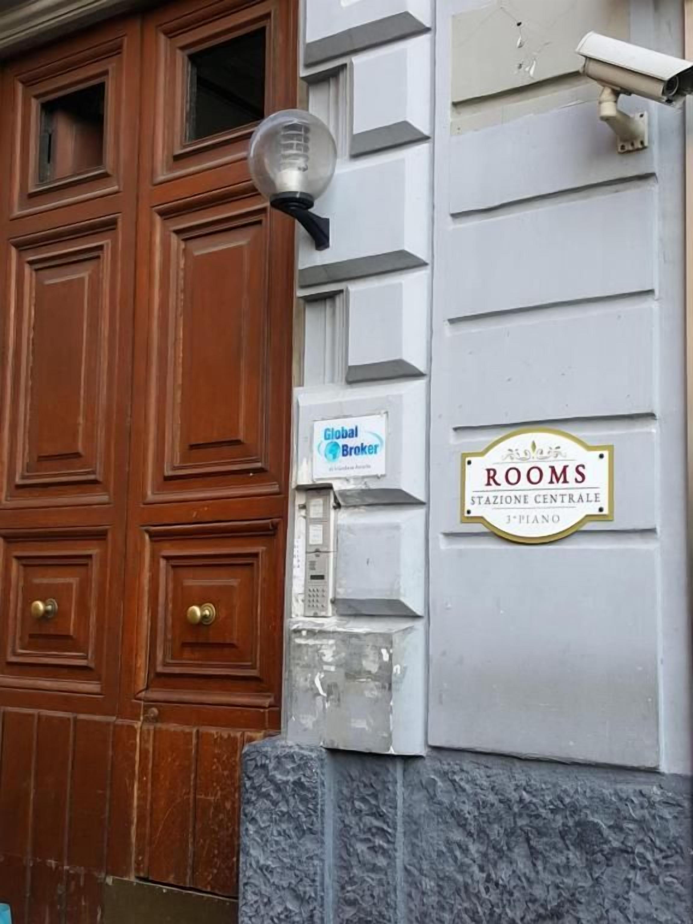 En byggnadsfasad med en trädörr, en gatlykta, en skylt med texten ”Global Broker Rooms” och en plakett som visar ”Stazione Centrale, 3:e våningen”.